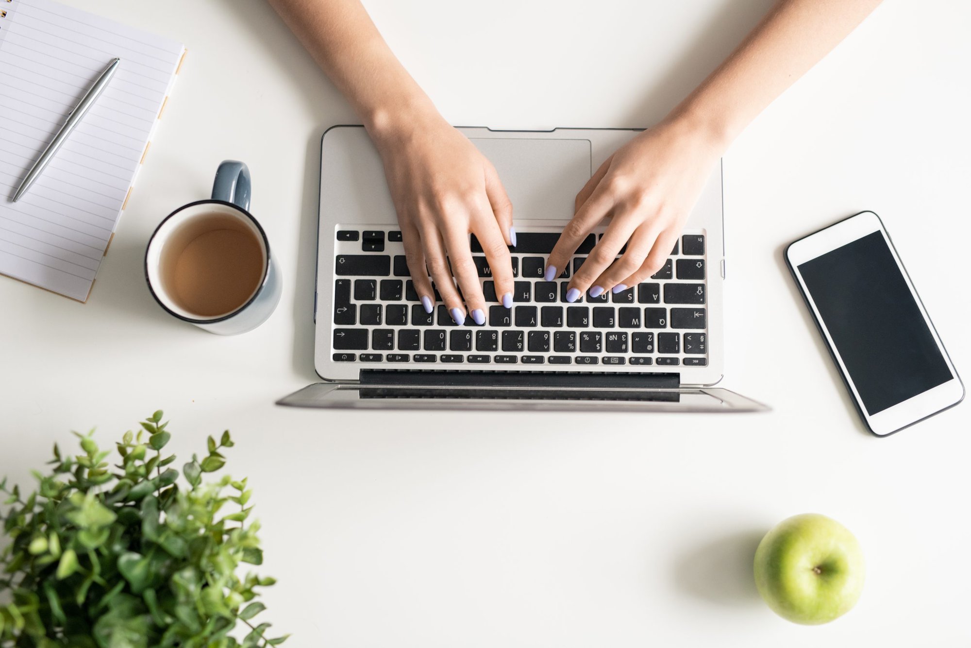 Top view of young female freelancer hands over keypad pf laptop during network with apple, mug of tea and smartphone near by